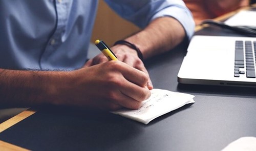 man writing on a notepad in front of a laptop.jpg