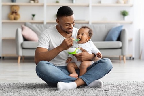 A young parent feeding their child while sat cross legged on the floor